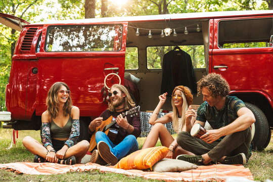 Group of friends hippies men and women laughing, and playing guitar near vintage minivan into the nature
