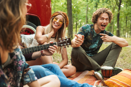 Group of smiling people hipsters men and women laughing, and sitting near vintage minivan in forest