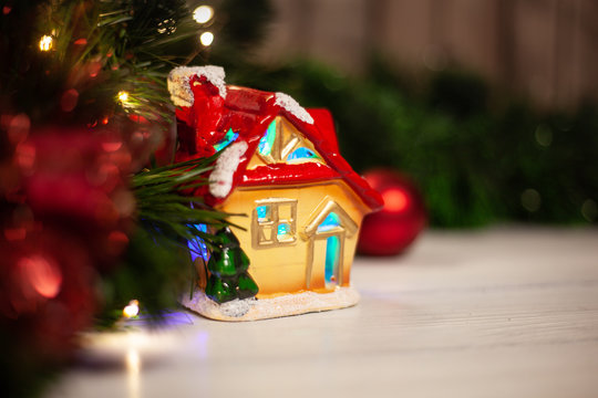 Christmas toy house with a red roof and windows with turquoise light next to a Christmas tree with a garland and red Christmas balls on a white wooden table