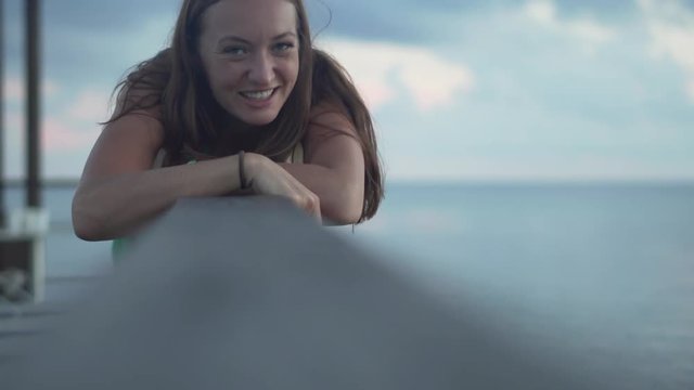 A woman is lying on the pier and is looking at the camera. She smiles and flirts. The ocean is near. The girl is on vacation. She is happy and calm