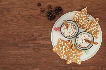 top view of cups with hot chocolate and marshmallows, cinnamon sticks and gingerbread cookies on wooden table