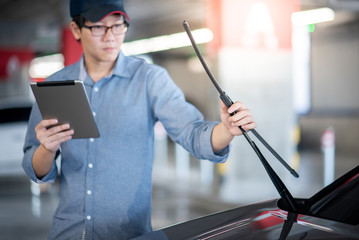 Young Asian auto mechanic holding digital tablet checking windshield wiper in auto service garage. Mechanical maintenance engineer working in automotive industry. Automobile servicing and repair - obrazy, fototapety, plakaty