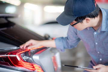 Young Asian auto mechanic holding digital tablet checking tail light in auto service garage. Mechanical maintenance engineer working in automotive industry. Automobile servicing and repair concept