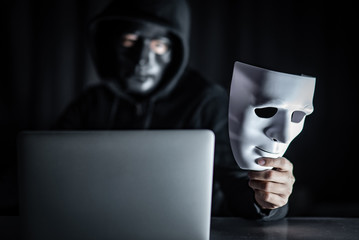 Mystery male hoodie hacker wearing black mask holding white mask with laptop computer on the table. Anonymous social masking. Ransomware cyber attack or internet security concepts
