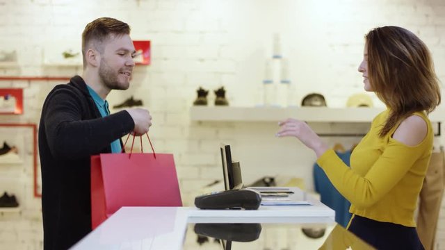 Cheerful customer is using a credit card to pay for purchased clothes while standing at cashier's desk. Shop assistant is accepting transaction and giving paper bags.