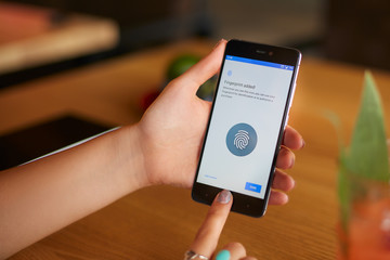 Female scanning fingerprint on her smartphone with program script on laptop display on backround. Woman unlock mobile phone with biometric sensor and finger. Privacy and security concept.