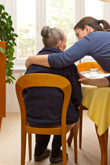 Rear view of a senior woman sitting at home beeing embraced by her granddaughter who is helping her during the day, eldercare by family members