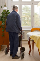 Rear view of an old woman moving around in her living room with 3-wheeled walker. A mobility device...