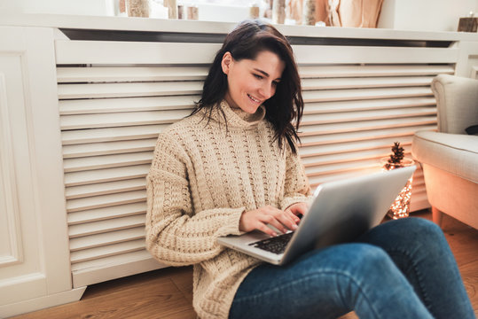 Image of young smiling woman shopping online on laptop in cozy christmas interior. Happy female in casual sitting on the floor, prepare gifts to family for xmas, bying on winter sales. Copy space.