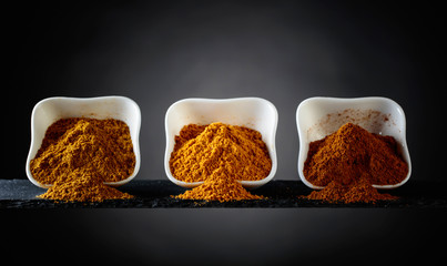 Small white ceramic bowls of Indian spices on dark background .
