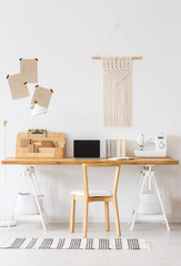 Real photo of a modern home offfice interior with a desk, laptop, sewing machine, chair and macrame on a wall. Empty screen, place your logo