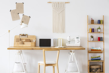 Real photo of a wooden desk with a laptop, sewing machine, organizer and macrame o a wall next to a shelf. Empty screen, place your logo