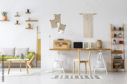 Real photo of a natural home office interior desk organizer, macrame on a wall, shelves and couch