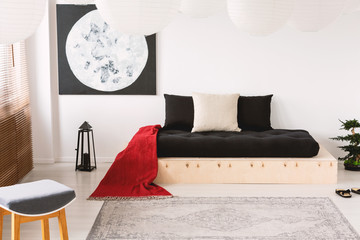 Red blanket on black wooden couch in white apartment interior with moon poster and stool. Real photo