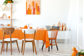 Real photo of kitchen table covered with orange tablecloth and white dishes. Artwork on the wall and shelf in the corner