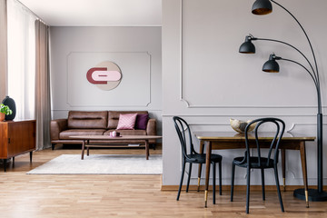 Black chairs at table next to lamp in white apartment interior with poster above sofa. Real photo