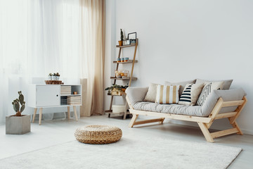 Cactus next to white cabinet in grey loft interior with pouf in front of wooden couch. Real photo