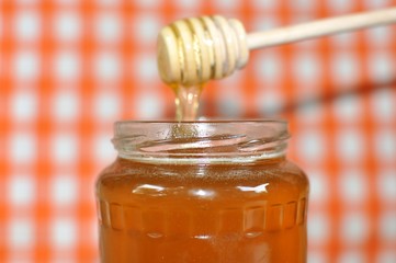 Pouring aromatic golden poly floral honey from wooden dipper into jar, on white and orange grid background, close up, selective focus