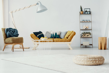 Pouf and chair in white living room interior with lamp next to mustard wooden sofa. Real photo
