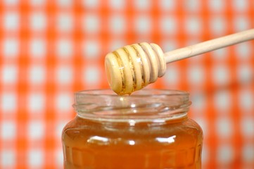 Pouring aromatic golden poly floral honey from wooden dipper into jar, on white and orange grid background, close up, selective focus