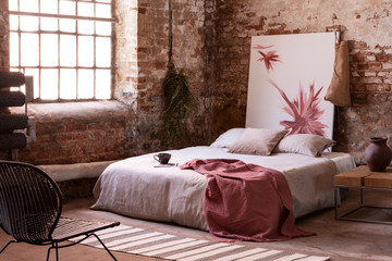 Red blanket and pillows on grey bed and chair in bedroom interior with poster and window. Real photo