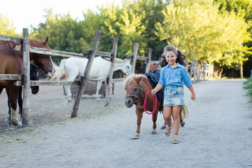 A small smiling girl with curly hair dressed in jeans walking with a pony at the stable