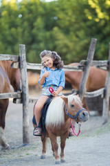 A small smiling girl with curly hair dressed in jeans riding a pony at the stable