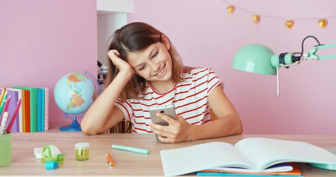 Cheerful teenage girl sitting at the desk and laughing, using cell phone. Dolly shot