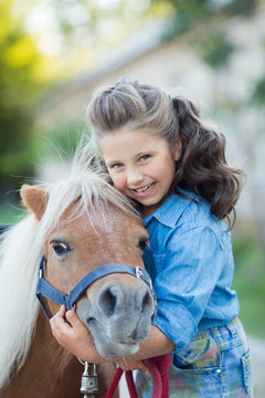 A small smiling girl with curly hair dressed in jeans walking with a pony at the stable
