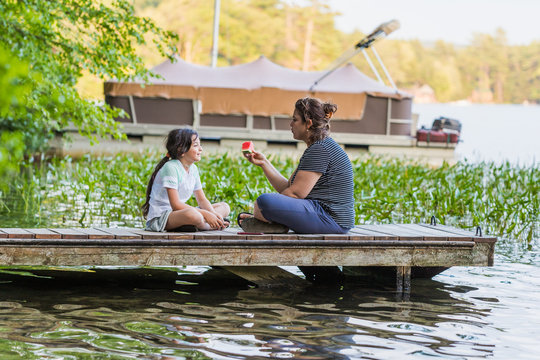 A woman is holding a piece of watermelon is talking to her young daughter while sitting on a dock