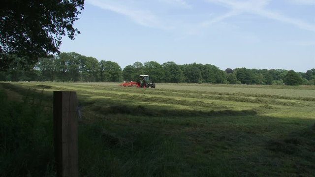 Farmer on tractor with rake attachment haying in grassland - collects cut hay into windrows for later collection.