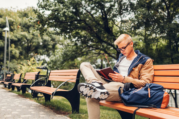 College student with backpack reading book walking in autumn park sitting on bench
