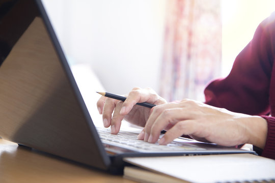 Hands Of A Man In Red Sweater Holding A Pencil While Typing On Laptop Computer Notebook Keyboard In The Office. Closeup With Left And Top Copy Space And Easy To Be Cropped As A Banner Or Strip.