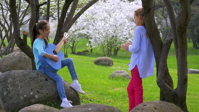 Two little girlfriend playing on jeans guitar and singing in city park, beautiful scenery