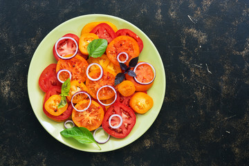 Red and yellow fresh tomato salad with onion, basil on a green plate, dark rustic background. Top view, copy space.