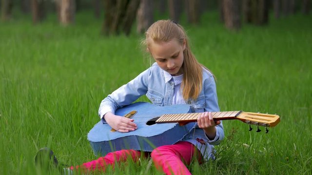 Young and beautiful little blonde girl sitting in green grass playing jeans guitar outdoor. spring season in countryside. Traveling concept.