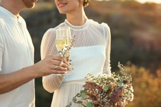 Happy newlyweds with beautiful field bouquet and glasses of champagne outdoors, closeup