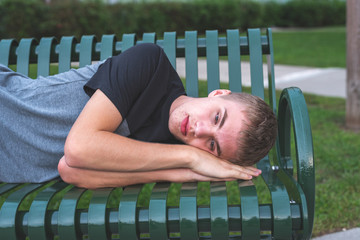Sad teenager lying down on an outdoor bench next to his high school.
