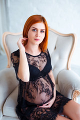 Fototapeta premium Pregnancy beauty. Beautiful elegant readhead pregnant woman in charming black dress posing in tender home interior.