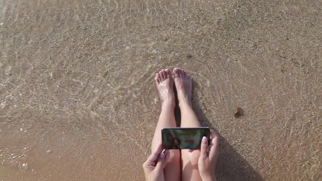 White beautiful tanned woman takes selfie while sitting at sunny morning beach using digital camera of modern smartphone to share in social media resources. Real time full hd video footage.