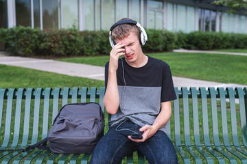 Depressed teenager sitting on bench outside of his school while listening to music with his headphones.