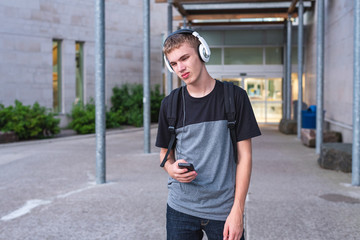 Sad teenager standing in front of his school while listening to music with his headphones.