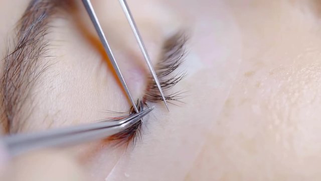 master of lash extension is putting false lashes near natural on eyelid of young woman, close-up