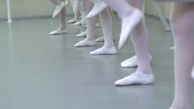 closeup legs of little ballerinas group in white shoes in row practicing in ballet studio slow motion. Young girls training elements of classical dance exercise. Childhood, dancing, lifestyle concept