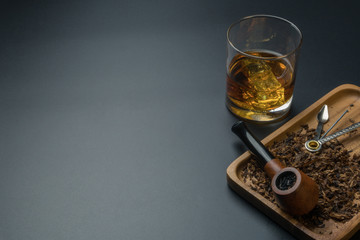 a smoking pipe with pipe tamper tool and tobacco in wooden tray, a glass of whiskey on the black table