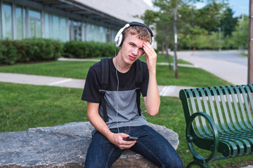 Fototapeta premium Ashamed teenager sitting on a rock outside of a school while listening to music.