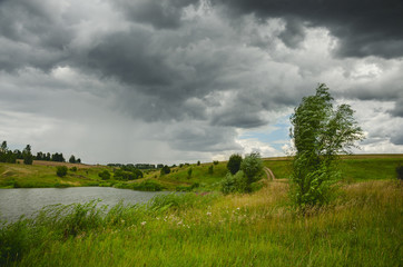 Cloudy summer landscape with small river.Dark stormy clouds in dramatic overcast sky.Fields,green meadows and woods.Rainy season.