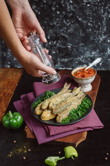 Chef's hands sprinkle with seasoning Fried fish in a plate with  tomato sauce, greens and peppers on wooden board  and a dark background