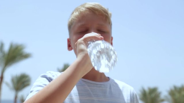 Closeup view of white european kid in casual clothes drinking fresh clear water from plastic bottle outdoors on hot sunny summer day. Focus at boottom of bottle, face of boy is blurry.