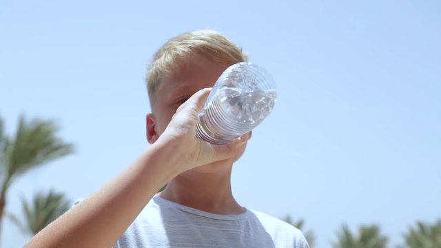 Closeup view of white european kid in casual clothes drinking fresh clear water from plastic bottle outdoors on hot sunny summer day. Boy isolated at blurry green palms and blue sky, sea background.